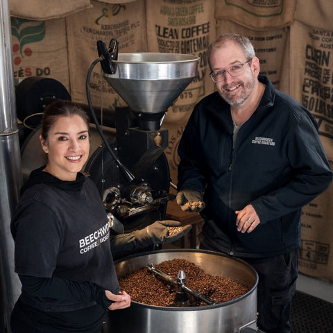 Karla and Angus smiling beside the coffee roaster, holding green beans over the cooling tray of freshly roasted coffee