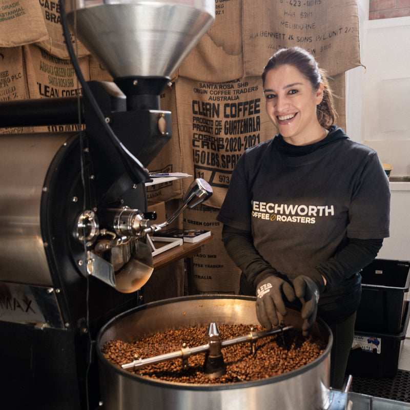 Karla smiling at the roaster beside the cooling tray of freshly roasted beans, with sacks of green coffee behind