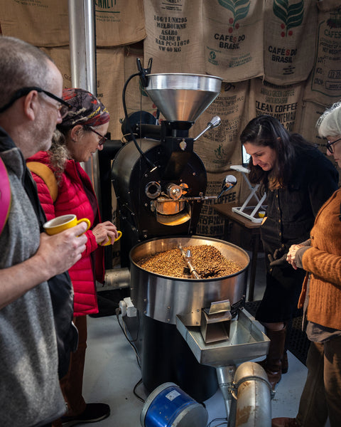Visitors with coffee in hand gather round the roaster as a fresh batch of beans cools, green-coffee sacks lining the wall behind